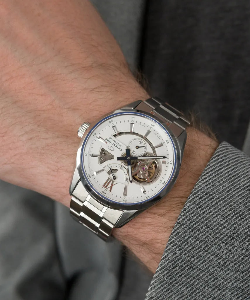 Primo piano di un polso che indossa l'Orient Star Skeleton Automatico RE-AV0113S00B di Orient, un raffinato orologio giapponese meccanico a vista con quadrante bianco e cinturino in acciaio inossidabile; la persona indossa una giacca grigia.
