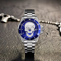 L'Orologio con teschio 3D silver blue di PACOSHOP, caratterizzato da un sorprendente design a teschio e da una lunetta blu, poggia su una superficie strutturata con una catena e una chiusura metalliche sfocate visibili sullo sfondo.
