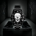 L'Orologio con teschio 3D All Black di PACOSHOP, caratterizzato da un cinturino in metallo e da un teschio argentato con un occhio di pietra preziosa sul quadrante, è esposto su un piedistallo di marmo nero su uno sfondo scuro.
