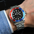 Un PACOSHOP24.COM Orologio automatico uomo meccanico mod. Gmt con lunetta rossa e blu, quadrante nero e bracciale in metallo, indossato al polso di una persona. Il quadrante dell'orologio riporta il marchio PACOSHOP24.COM e la dicitura "Automatic".