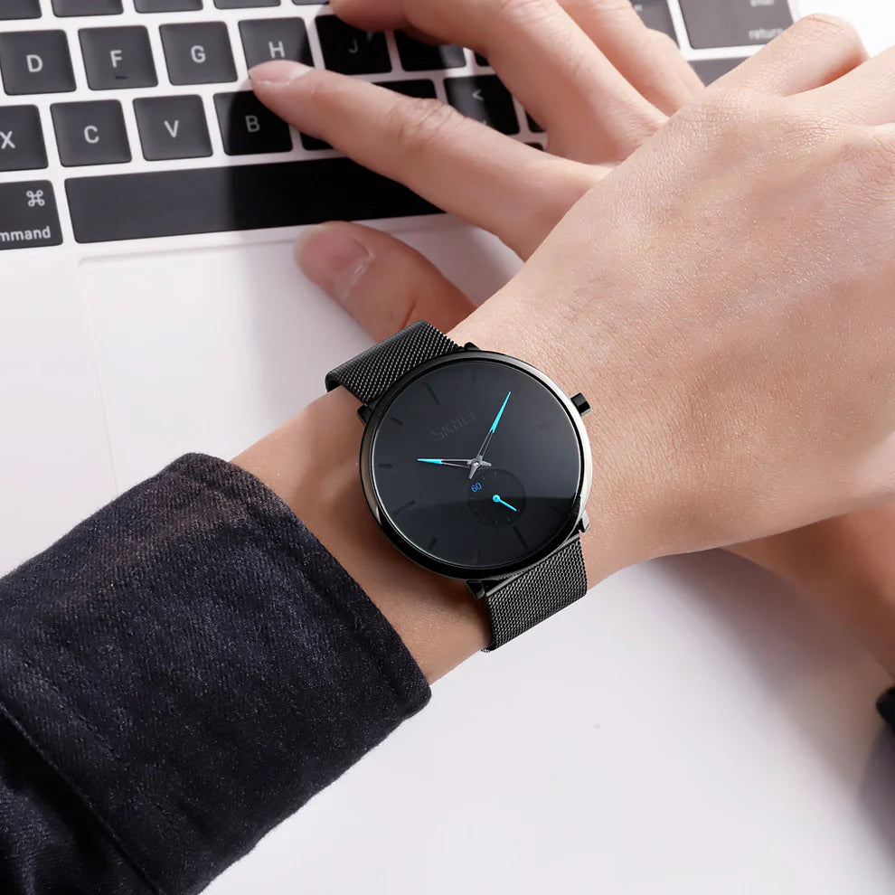 Una persona che indossa lo Skmei Orologio minimalista ultraleggero ultrasottile nero blu con cinturino a rete nero e quadrante nero con lancette blu digita su un computer portatile.