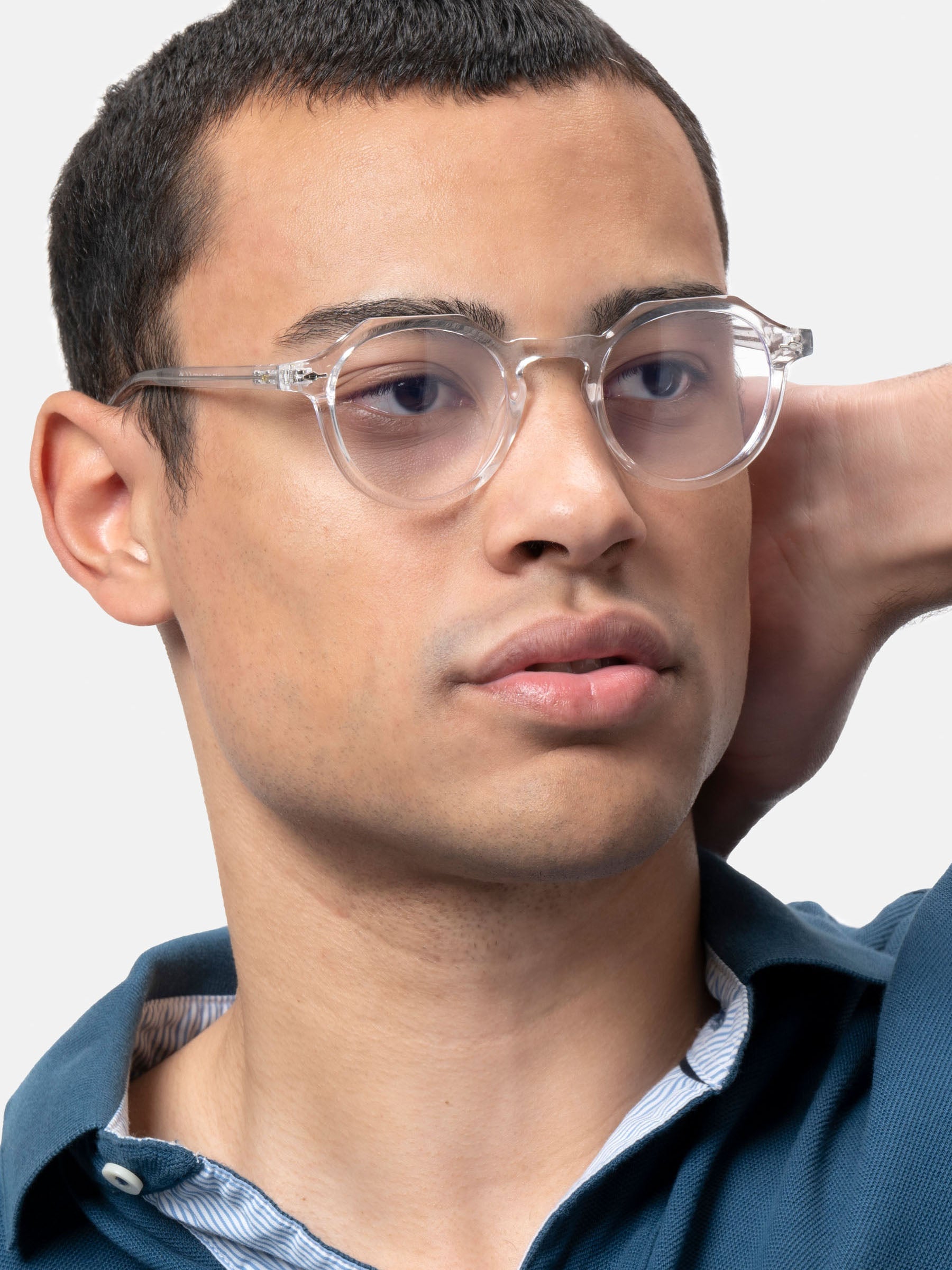 Un giovane uomo in camicia blu indossa X Frame Therapy Occhiali luce blu Lucentia lenti trasparenti Unisex e guarda pensieroso di lato con una mano dietro la testa su uno sfondo chiaro.