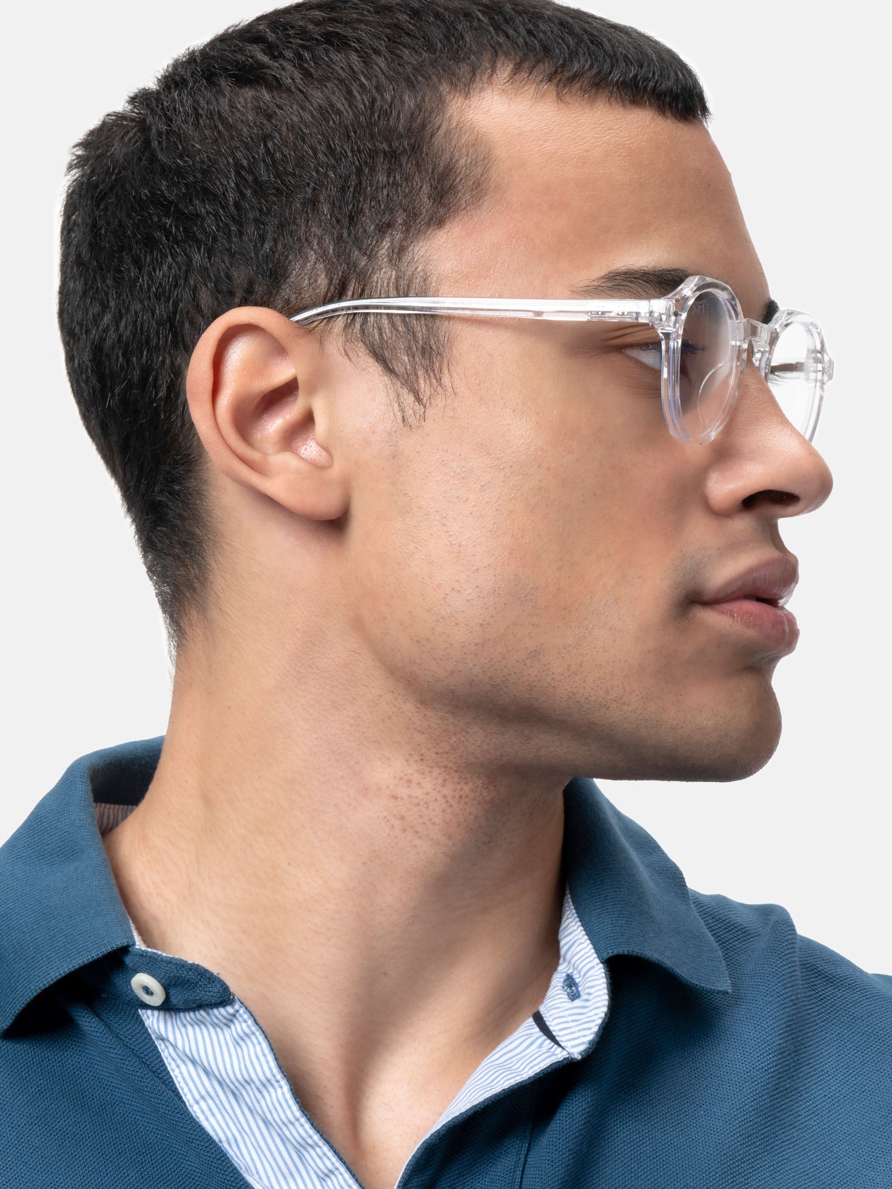 Un giovane uomo con i capelli corti e scuri indossa X Frame Therapy Occhiali luce blu Lucentia lenti trasparenti Unisex e una camicia con colletto blu, guardando di lato su uno sfondo bianco.