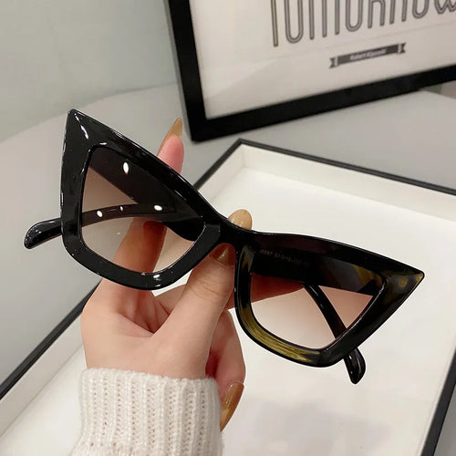 Una mano con unghie lunghe e curate regge PACOSHOP24.COM Occhiali da Sole Donna Occhi di Gatto - Fashion Style. Lenti colorate e un'opera d'arte incorniciata sullo sfondo sottolineano l'eleganza di questi occhiali da sole cat-eye.