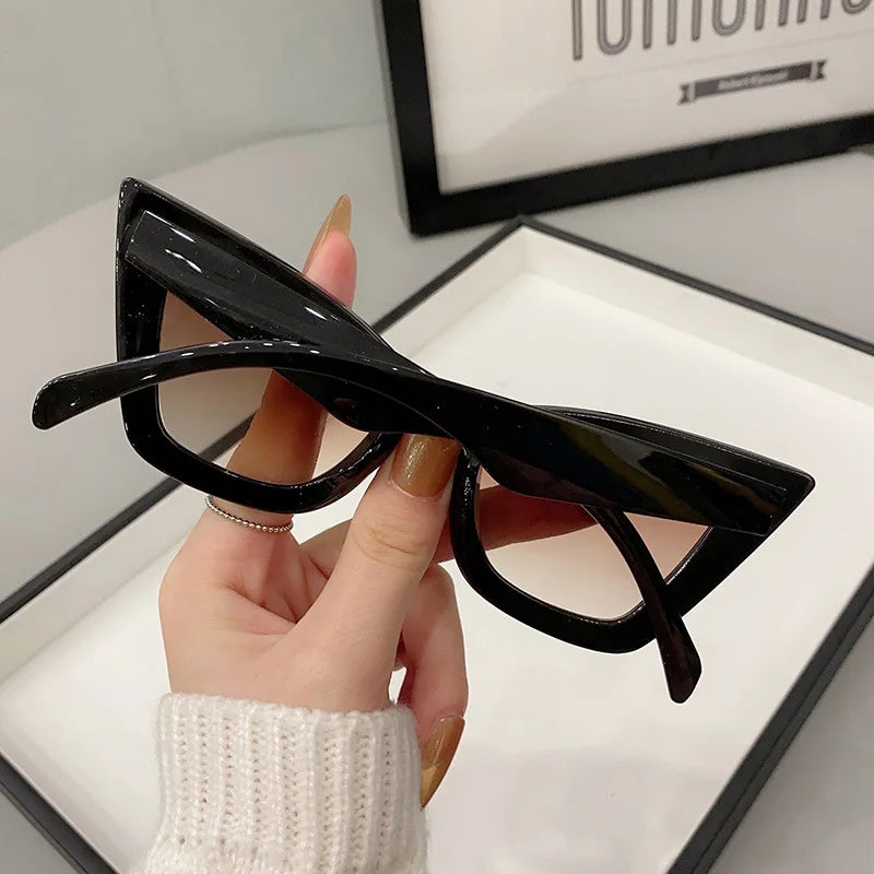 Una mano con unghie lunghe e curate tiene un paio di PACOSHOP24.COM Occhiali da Sole Donna Occhi di Gatto - Fashion Style davanti a un vassoio bianco e a una foto incorniciata.