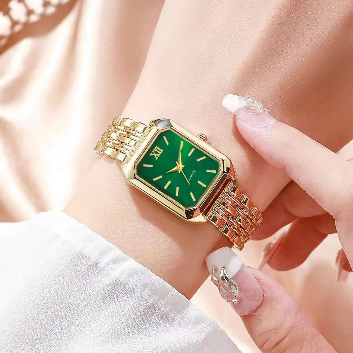 Una mano indossa un PACOSHOP24.COM Orologio al quarzo donna rettangolare alla moda con cinturino in oro, quadrante verde e indici in oro. Un'altra mano con unghie lunghe e decorate tocca delicatamente l'elegante orologio; lo sfondo è leggermente sfocato.