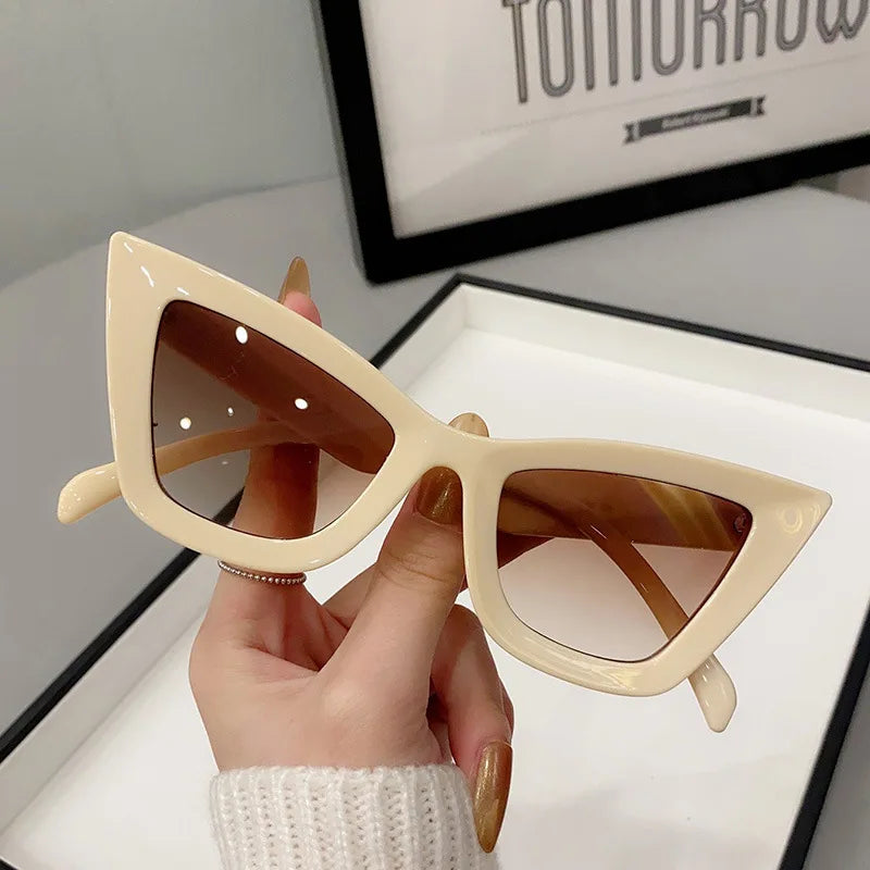 Una mano con unghie lunghe e curate regge gli Occhiali da Sole Donna Occhi di Gatto - Fashion Style di PACOSHOP24.COM: occhiali da sole cat-eye chic color crema con lenti marroni. Sullo sfondo si intravede parzialmente una stampa incorniciata.