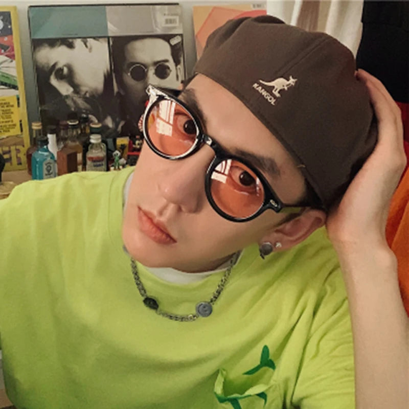 Una persona che indossa PACOSHOP24.COM Occhiali da Sole Unisex stile Ovale, un berretto Kangol marrone, una camicia verde neon e una collana a catena posa con una mano sulla testa. Sullo sfondo compaiono bottiglie e poster.