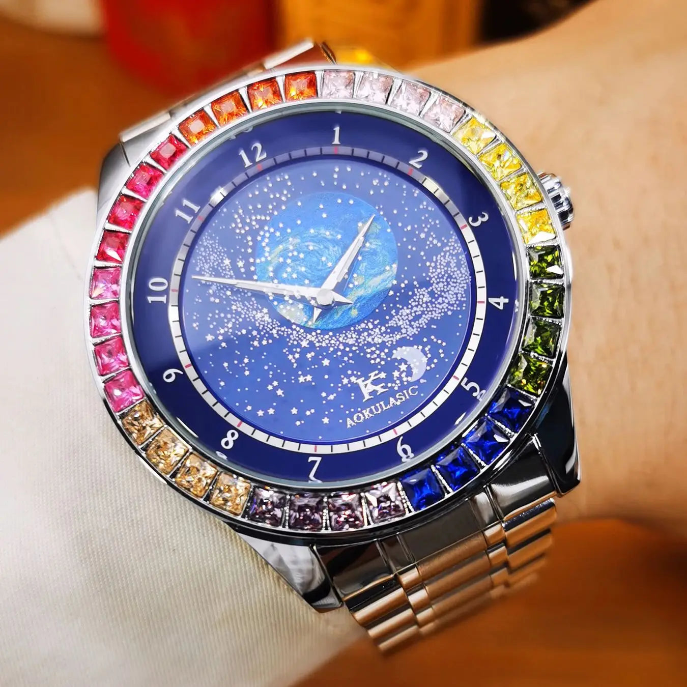 L'Orologio automatico uomo cielo stellato Design luminoso di PACOSHOP24.COM presenta una cassa argentata, un quadrante blu a tema stellare e una lunetta ornata di gemme sfumate arcobaleno per un look lussuoso. Mostrato al polso con una guaina chiara.