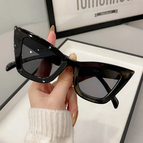Una mano con le unghie lunghe e un maglione bianco tiene in mano PACOSHOP24.COM Occhiali da Sole Donna Occhi di Gatto - Fashion Style, con lenti scure, su una superficie bianca con una foto incorniciata sullo sfondo: un tocco chic di eleganza cat-eye.