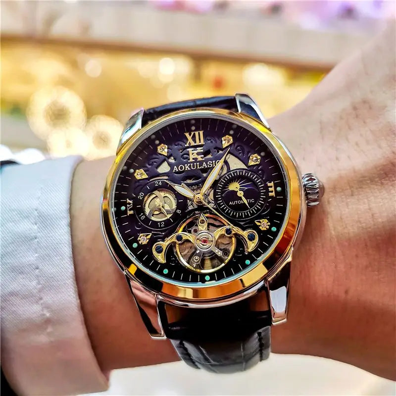Primo piano di un polso che indossa un PACOSHOP24.COM Orologio automatico uomo con fasi lunari, con quadrante nero, ingranaggi a vista, numeri romani e cinturino in pelle nera. Lo sfondo è sfocato da un'illuminazione calda.