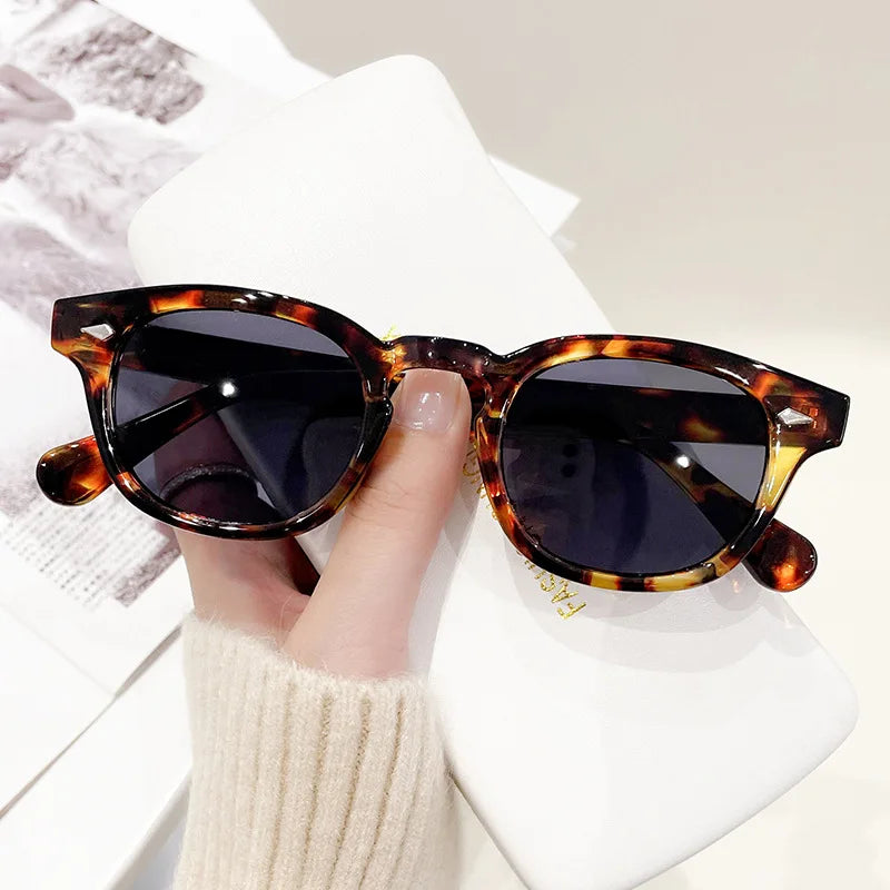 Una mano regge PACOSHOP24.COM Occhiali da sole vintage fashion retrò rotondi Unisex Policarbonato con lenti scure sopra una custodia bianca. La persona, in maglione chiaro, ha una rivista o un giornale parzialmente visibile sullo sfondo.