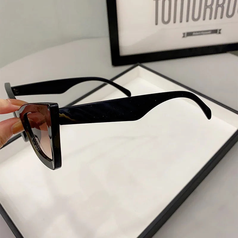 Una mano regge PACOSHOP24.COM Occhiali da Sole Donna Occhi di Gatto - Fashion Style, occhiali da sole neri con montatura spessa e lenti scure, visti di lato su una superficie bianca con una stampa a cornice parzialmente visibile.