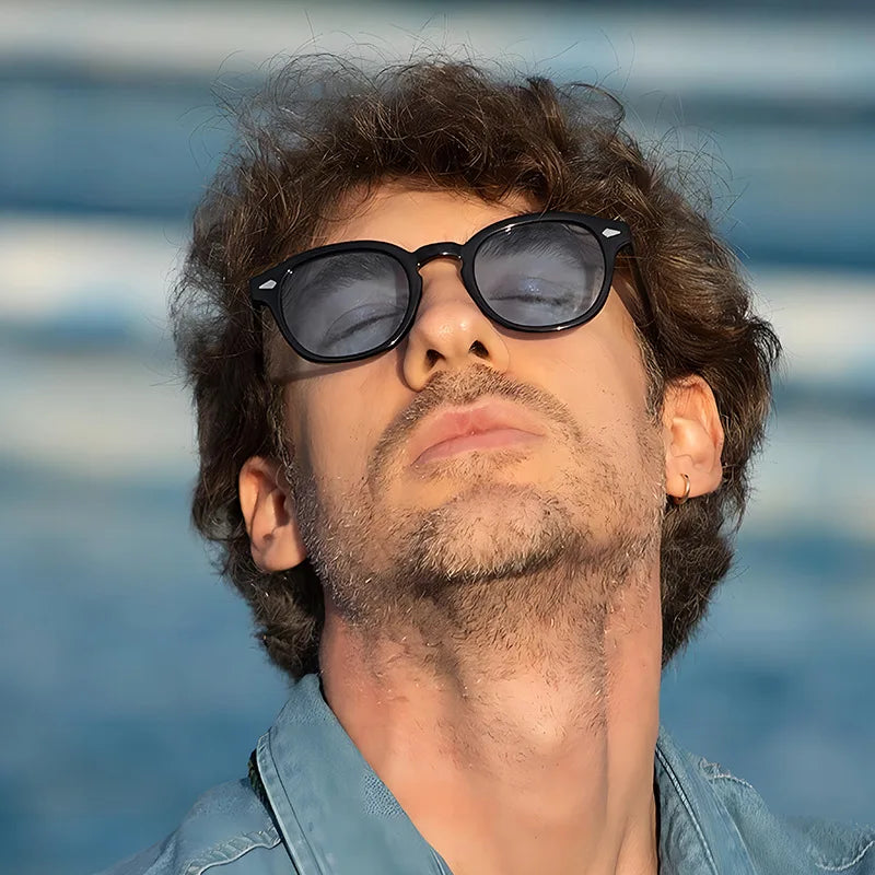 Una persona con i capelli ricci indossa PACOSHOP24.COM Occhiali da sole vintage fashion retrò rotondi Unisex Policarbonato, inclinando la testa all'indietro e guardando verso l'alto su uno sfondo sfocato bianco e blu che suggerisce acqua o onde.