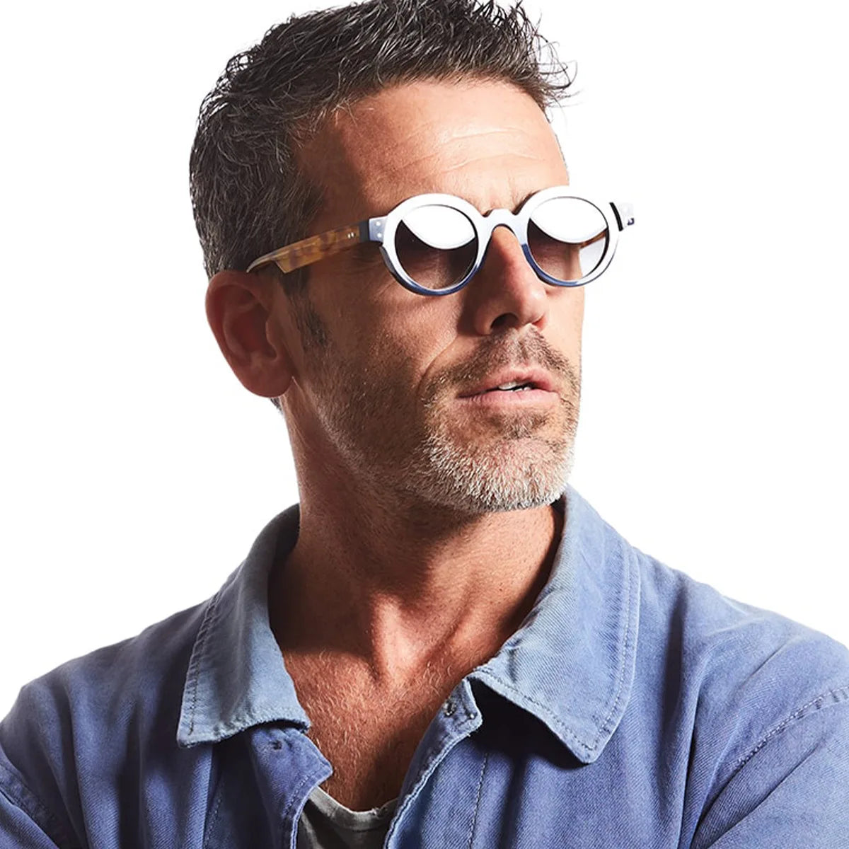 Un uomo con capelli corti, brizzolati e barbetta indossa PACOSHOP24.COM Occhiali da Sole Rotondi punk Retrò UV400 - Uomo Donna con montatura bianca e camicia con colletto blu, guardando leggermente di lato su uno sfondo bianco.