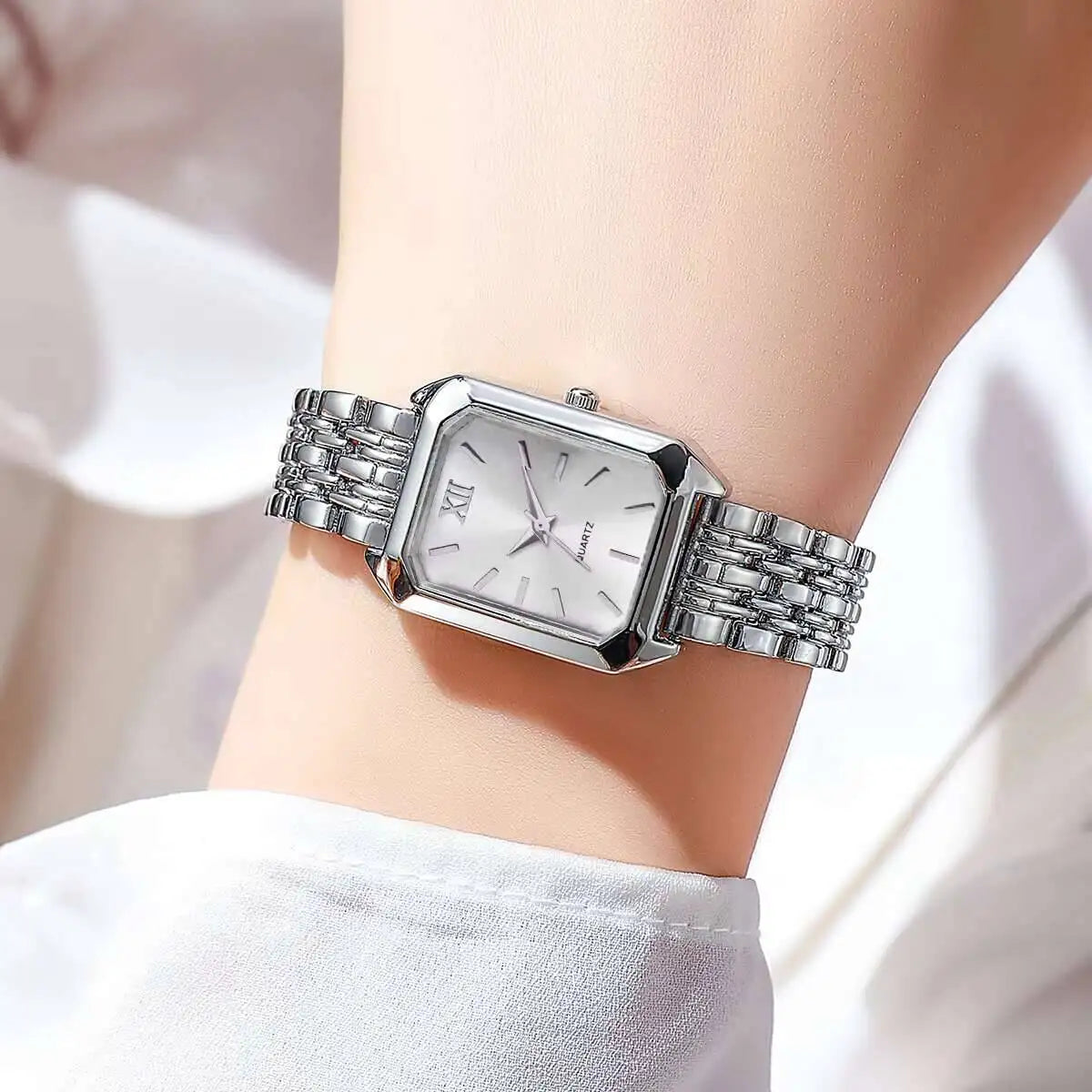 Una donna indossa l'orologio PACOSHOP24.COM al quarzo donna rettangolare alla moda & Fashion - argento con quadrante bianco, numeri romani e fascia a maglie in metallo - che si staglia elegantemente su uno sfondo illuminato in modo soffuso.