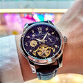 Primo piano di un polso che indossa un PACOSHOP24.COM Orologio automatico uomo con fasi lunari, con cinturino in pelle nera, ingranaggi visibili e indici a numeri romani. Lo sfondo sfocato si illumina di luci colorate.