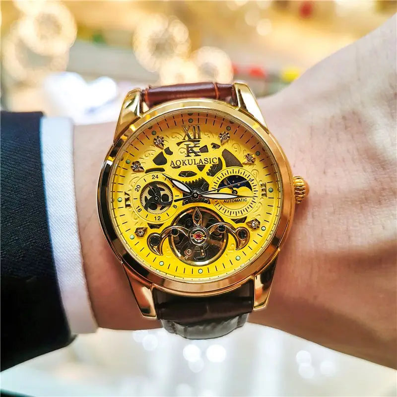 Primo piano del polso di una persona che indossa un PACOSHOP24.COM Orologio automatico uomo con fasi lunari, con quadrante scheletrato in oro, cinturino in pelle marrone e ingranaggi a vista, su uno sfondo delicatamente sfocato e illuminato in modo caldo.