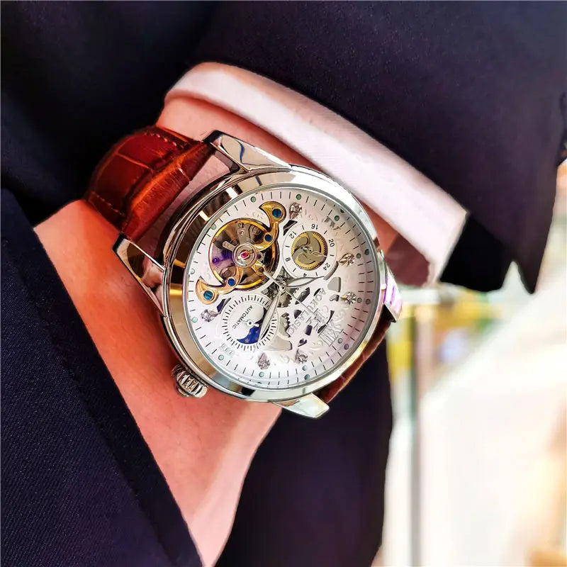 Una persona in giacca e cravatta indossa un PACOSHOP24.COM Orologio automatico uomo con fasi lunari, con quadrante scheletrato argentato, ingranaggi a vista e cinturino in pelle marrone che ne evidenzia gli intricati dettagli meccanici.