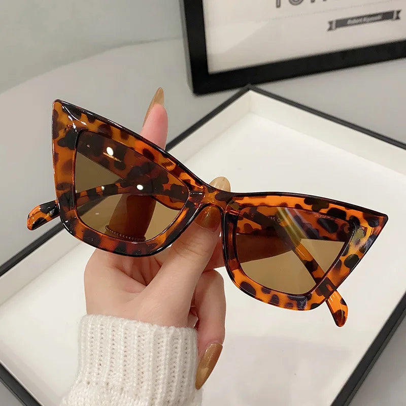 Una mano con unghie lunghe e curate sorregge gli Occhiali da Sole Donna Occhi di Gatto - Fashion Style di PACOSHOP24.COM, caratterizzati da una montatura marrone tartarugata e da lenti colorate, su un nitido sfondo bianco.