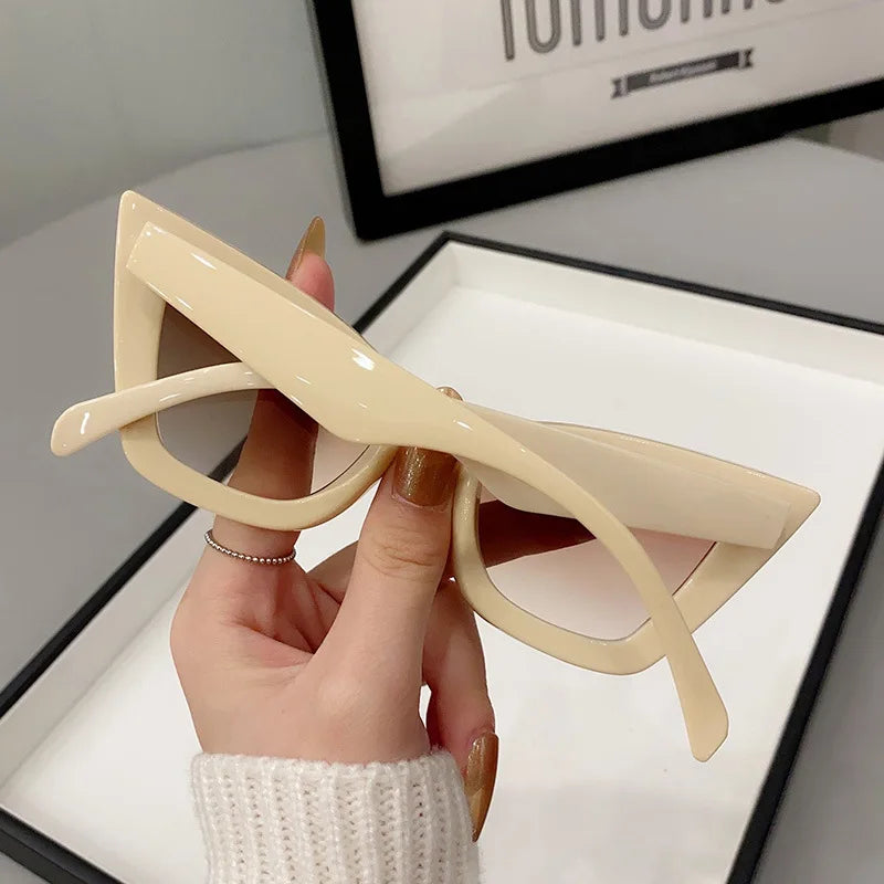 Una mano con le unghie dipinte regge gli Occhiali da Sole Donna Occhi di Gatto - Fashion Style di PACOSHOP24.COM. Un vassoio bianco e un'insegna incorniciata parzialmente visibile completano un look chic e di tendenza.