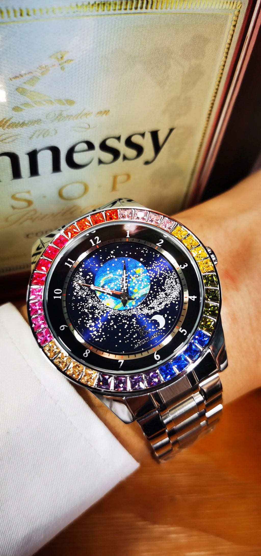 Un polso impreziosito dall'Orologio automatico PACOSHOP24.COM uomo cielo stellato Design luminoso, con lunetta tempestata di gemme e quadrante cielo stellato, con fasi lunari e dettagli terrestri, in posa davanti a una bottiglia Hennessy V.S.O.P.