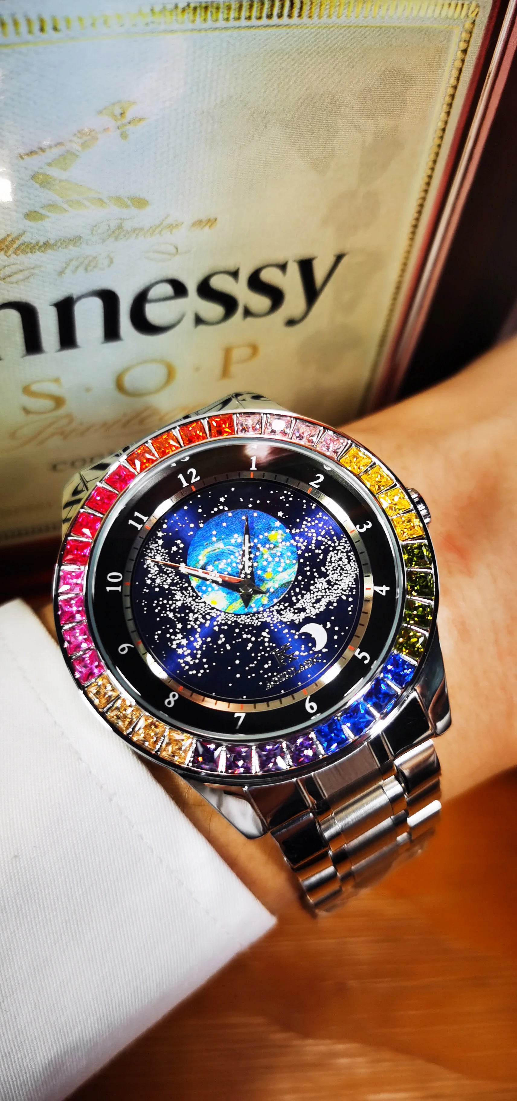 Un polso impreziosito dall'Orologio automatico PACOSHOP24.COM uomo cielo stellato Design luminoso, con lunetta tempestata di gemme e quadrante cielo stellato, con fasi lunari e dettagli terrestri, in posa davanti a una bottiglia Hennessy V.S.O.P.