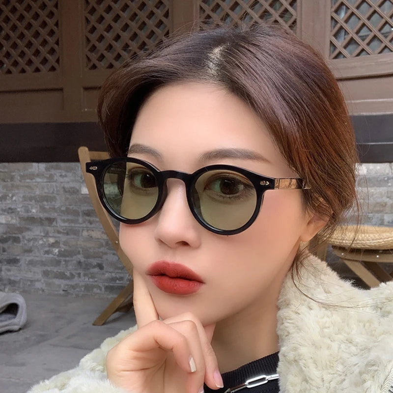 Una donna con i capelli scuri indossa PACOSHOP24.COM Occhiali da Sole Unisex stile Ovale moda - occhiali da sole neri rotondi con lenti UV400 - e un cappotto di pelliccia leggera, in posa all'aperto accanto a un muro di mattoni e a una sedia di legno.