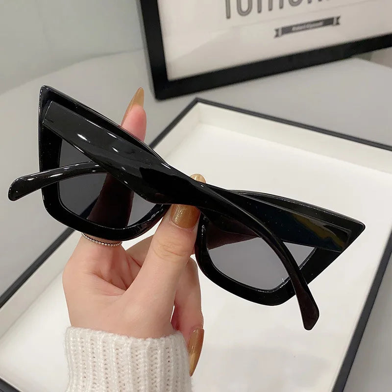 Una mano con unghie lunghe e curate tiene in mano gli Occhiali da Sole Donna Occhi di Gatto - Fashion Style di PACOSHOP24.COM in casa, con un vassoio bianco e un oggetto incorniciato sullo sfondo.
