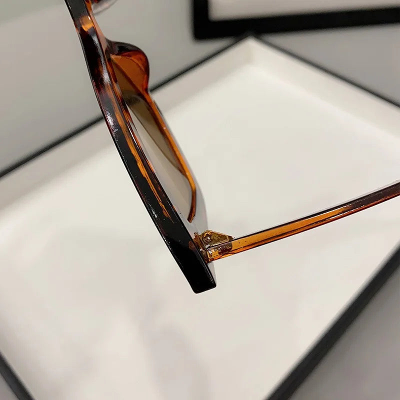 Primo piano di PACOSHOP24.COM Occhiali da Sole Donna Occhi di Gatto - Fashion Style in tartaruga con lenti marroni, che mostra il lato e la cerniera, appoggiato su una superficie bianca con una cornice nera sullo sfondo.