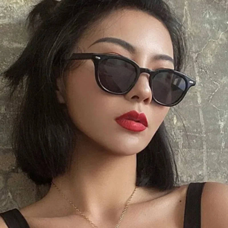 Una donna con i capelli corti neri indossa Occhiali da Sole Unisex stile Ovale moda di PACOSHOP24.COM e un rossetto rosso brillante. Abbina un top nero a una collana d'oro e posa con stile contro una parete grigia.