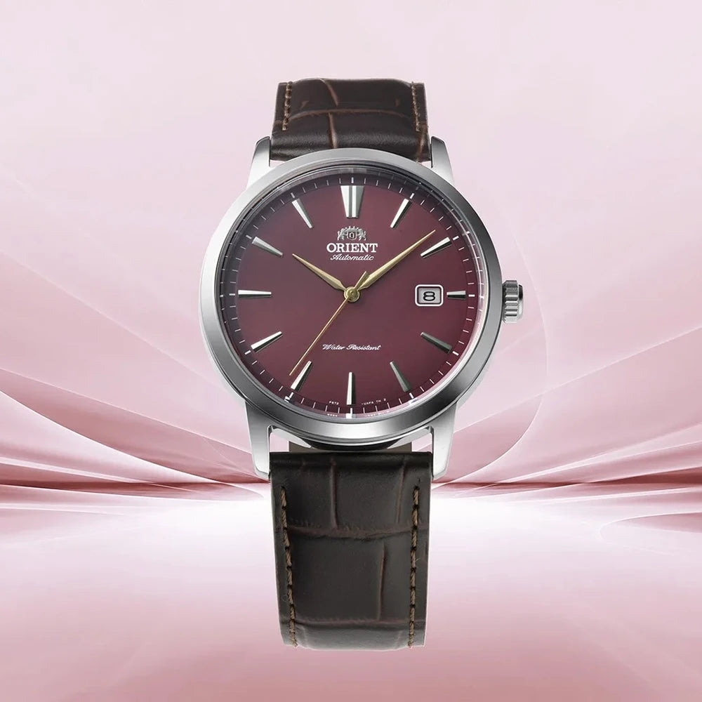 L'Orient Bambino RA-AC0F15R30B di Orient è un orologio automatico da uomo con un sorprendente quadrante rosso, indici argentati, datario a ore 3, cinturino in pelle marrone e cassa in acciaio inossidabile su uno sfondo astratto rosa tenue.