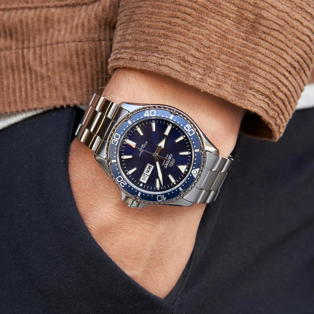 Una persona con una giacca di velluto a coste marrone tiene la mano sinistra in tasca e mostra un Orient Mako III Automatico RA-AA0002L39B, un orologio subacqueo automatico da uomo con lunetta blu e quadrante nero che indica giorno e data.