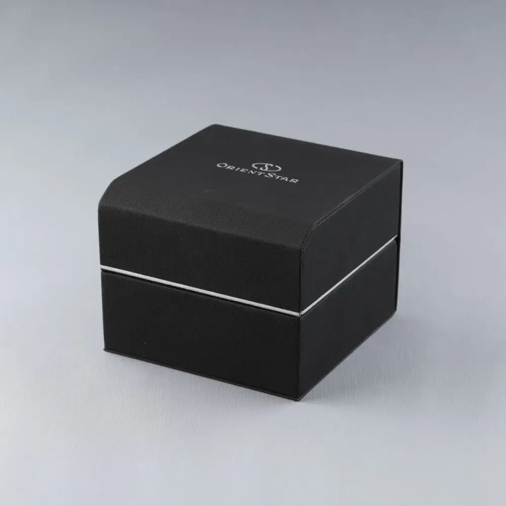 Una scatola nera e quadrata di Orient Star, con una striscia argentata e il logo bianco sulla parte superiore, si trova su una superficie grigio chiaro, ideale per l'Orient Star Classic RE-AU0109L00B, un elegante orologio automatico per uomo e donna di Orient.