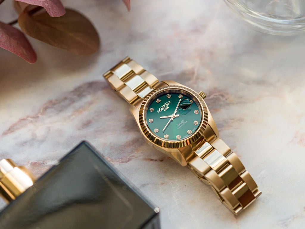 Un Roamer Amalfi 852844487920 orologio donna d'oro con quadrante verde e indici di diamanti poggia su un marmo vicino a una bottiglia di profumo nera e a foglie marroni, dimostrando uno stile raffinato.