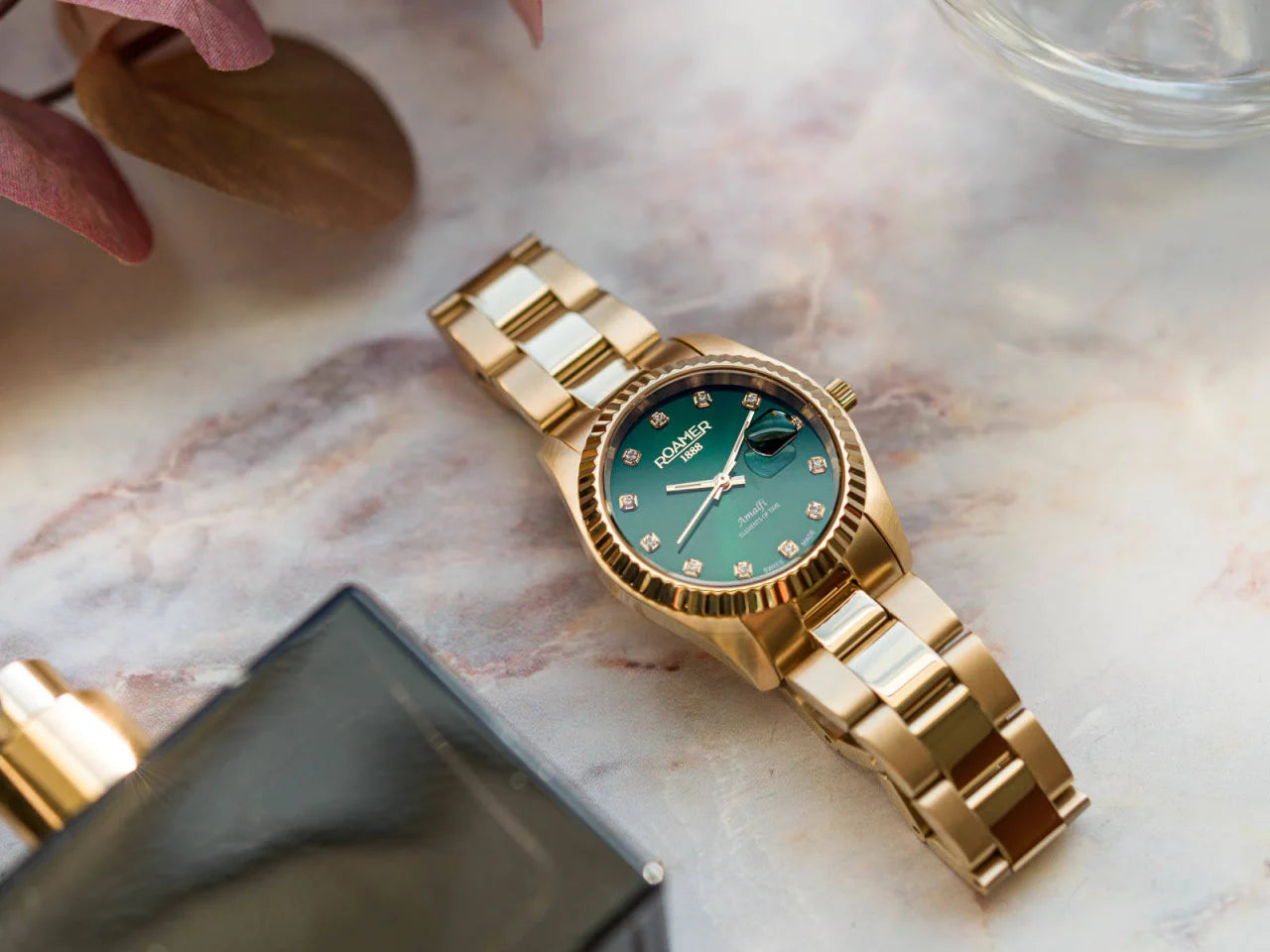Un Roamer Amalfi 852844487920 orologio donna d'oro con quadrante verde e indici di diamanti poggia su un marmo vicino a una bottiglia di profumo nera e a foglie marroni, dimostrando uno stile raffinato.
